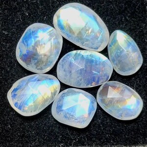 May include: Seven faceted moonstone gemstones with a pearlescent sheen. The stones display a range of shapes, including oval and teardrop, and reflect a spectrum of colors. The gemstones are set against a dark background.