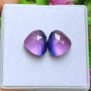 Puede incluir: Dos gemas facetadas con un degradado de tonos morados y azules. Las gemas tienen forma de corazón y están sobre una superficie blanca. Las gemas son probablemente para la fabricación de joyas.