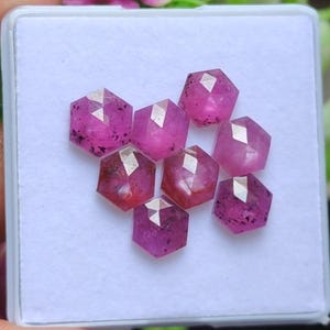 Puede incluir: Siete rubíes hexagonales facetados en una caja blanca. Las piedras preciosas son de un rosa vibrante con inclusiones más oscuras. Cada piedra tiene múltiples facetas que reflejan la luz, creando un efecto brillante. Los rubíes están dispuestos en un grupo.