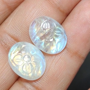 May include: Two oval-shaped, iridescent moonstone cabochons. Each stone is carved with a floral design. The stones display a range of colors, including blue, yellow, and white. The stones are held in a hand.
