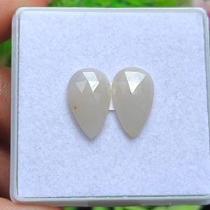 Op de afbeelding: Twee peervormige, gefacetteerde edelstenen met een bleke, crèmekleurige tint worden getoond in een witte doos. De edelstenen hebben een gepolijste, doorschijnende uitstraling, met meerdere facetten die het licht vangen. De setting is een eenvoudige, neutrale achtergrond, die de vorm en kleur van de stenen benadrukt.