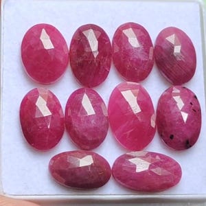 Puede incluir: Colección de nueve piedras preciosas de rubí ovaladas y facetadas. Los rubíes son de un color rojo intenso y vibrante, dispuestos en una bandeja blanca. Cada piedra tiene múltiples facetas que capturan la luz, creando un efecto brillante. Ideales para joyería.