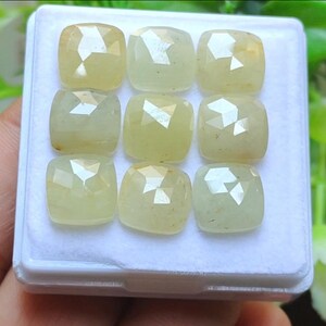 May include: Nine pale yellow gemstones with a cushion cut are displayed in a white tray. The gems have a faceted surface, reflecting light. The gems are likely for jewelry making.
