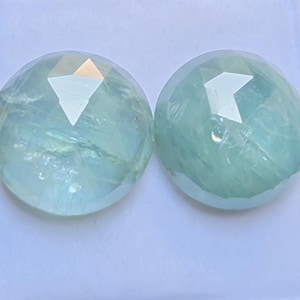 May include: Two round, faceted gemstones with a pale, translucent mint-green hue. Each stone features a polished, multi-faceted surface that reflects light, showcasing internal inclusions and a clear, glassy texture throughout the circular shape.