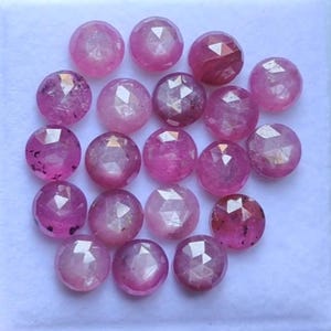 Puede incluir: Una colección de gemas redondas y facetadas en varios tonos de rosa y magenta. Las gemas tienen una superficie pulida y reflectante, con inclusiones internas visibles. La disposición es dispersa, mostrando las características individuales de las gemas.