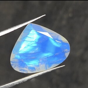 May include: A faceted, pear-shaped moonstone gemstone displaying a translucent white base with a vibrant, glowing blue adularescence across its surface, held securely by a pair of thin, silver-toned metal tweezers against a dark background.