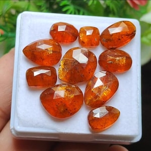 May include: A collection of faceted, orange-brown gemstones of varying shapes, including oval, square, and teardrop, are displayed in a white tray. The gems have a polished, reflective surface, showcasing their internal structure and color variations. The gems are likely for jewelry making.
