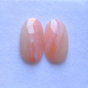 Puede incluir: Dos piedras preciosas ovaladas con una parte superior facetada. Las piedras son de color melocotón y crema, con remolinos e inclusiones internas. Las piedras preciosas están sobre un fondo blanco.