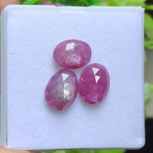 Puede incluir: Tres gemas rosas facetadas, de forma ovalada, se muestran sobre una superficie blanca. Las gemas tienen una superficie pulida y reflectante, y están dispuestas en un patrón triangular. Las piedras preciosas son de un tono rosa vibrante.