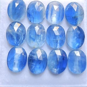 May include: A collection of twelve oval-shaped blue gemstones arranged in a four-by-three grid. Each stone features a faceted top surface with a smooth, rounded profile, displaying varying shades of translucent blue with internal inclusions.