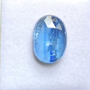 Puede incluir: Una piedra preciosa azul de forma ovalada y facetada, con apariencia translúcida e inclusiones internas visibles. La piedra muestra tonos variables de azul claro a medio con una superficie pulida y un corte geométrico distintivo.