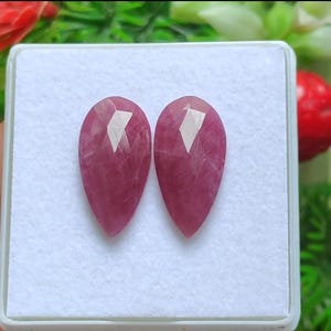 May include: Two faceted, teardrop-shaped gemstones in a white box. The gems are a deep pinkish-red color with internal veining and a polished, reflective surface. The gems are displayed against a white background.