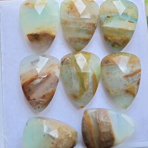 Puede incluir: Un conjunto de ocho piedras preciosas facetadas en forma de pera, con una base de color verde menta pálido e inclusiones de matriz en tonos marrón y bronceado. Cada piedra presenta una superficie pulida y multifacetada que refleja la luz.