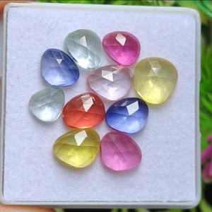 Könnte beinhalten: Eine Sammlung facettierter Edelsteine in verschiedenen Farben, darunter Blau, Rosa, Gelb und Klar. Die tropfenförmigen Edelsteine werden in einem weißen Kunststoffbehälter präsentiert. Die Edelsteine haben eine polierte, reflektierende Oberfläche.
