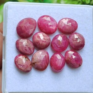 Op de afbeelding: Een verzameling van elf gefacetteerde, ovaalvormige edelstenen in verschillende tinten roze en rood. De edelstenen worden gepresenteerd in een witte, rechthoekige container. Elke steen heeft een uniek patroon en slijpsel, dat licht reflecteert.
