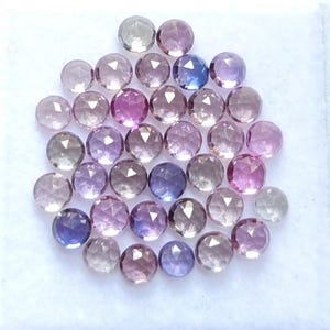 May include: A collection of small, round, faceted gemstones in a variety of colors including shades of pink, purple, blue, and clear white. Each stone features a geometric cut with multiple triangular facets that reflect light across the surface.
