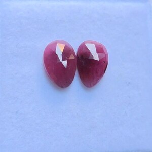 Peut inclure: Deux pierres précieuses en forme de goutte d'eau, facettées. Les gemmes sont d'un magenta profond avec de multiples facettes qui réfléchissent la lumière. Les pierres sont présentées sur une surface blanche, mettant en valeur leur aspect poli et leur teinte vibrante.