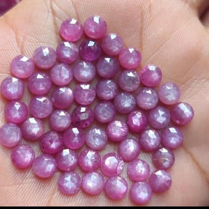 Könnte beinhalten: Eine Ansammlung facettierter, runder, rosa Edelsteine. Die Edelsteine haben eine polierte, reflektierende Oberfläche und sind eng beieinander angeordnet. Die Farbe variiert von hell- bis tiefrosa, wobei einige Steine durchscheinender erscheinen. Wahrscheinlich Rubine.