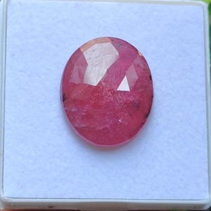 May include: Oval-shaped, faceted ruby gemstone. The gem is a deep pinkish-red color, with visible internal inclusions and a polished surface. The ruby is set against a white background within a square container.