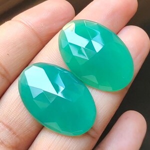 May include: Two oval-shaped green gemstones with a faceted cut. The gemstones are smooth and polished.