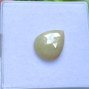 Op de afbeelding: Een peervormige, lichtgele edelsteen met een facetgeslepen oppervlak. De edelsteen is geplaatst tegen een witte achtergrond in een kleine, vierkante doos. De steen heeft een doorschijnende kwaliteit en reflecteert licht.