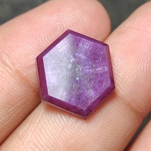 Peut inclure: Une pierre précieuse hexagonale facettée d'un violet profond. La gemme a une surface polie avec des veines internes et une zone centrale plus claire. Les bords sont bien définis, mettant en valeur la forme géométrique de la gemme. La pierre est tenue dans une main.