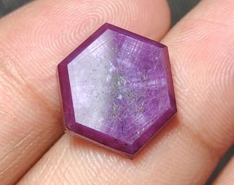 Puede incluir: Una gema facetada hexagonal con un tono púrpura intenso. La gema tiene una superficie pulida con vetas internas y un área central más clara. Los bordes están bien definidos, mostrando la forma geométrica de la gema. La gema se sostiene en una mano.