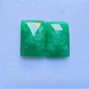 Può includere: Due pietre preziose di smeraldo rettangolari e sfaccettate. Le gemme sono di un verde vibrante con una superficie lucida e un taglio geometrico. Le sfaccettature riflettono la luce, creando un effetto scintillante. Le pietre sono su sfondo azzurro.