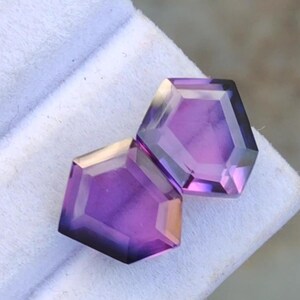 May include: Two faceted, hexagonal gemstones with a deep purple hue. The gems have a polished surface and a geometric design. The stones are set against a white background, highlighting their color and clarity. These gems are suitable for jewelry making.