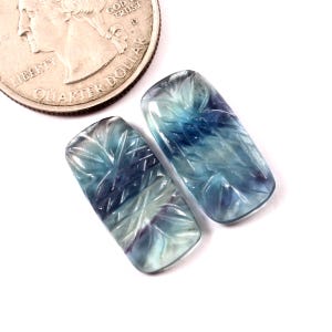 Op de afbeelding: Twee gegraveerde fluoriet kralen, één blauwe en één groene, met een gegraveerd bloemenmotief. De kralen zijn rechthoekig en hebben een gladde, gepolijste afwerking.