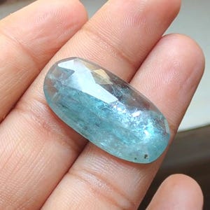 May include: A light blue aquamarine gemstone with a faceted oval shape. The gemstone is held in a hand.