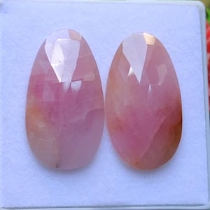 Op de afbeelding: Twee ovale, gefacetteerde edelstenen in roze tinten. De edelstenen hebben een gepolijst oppervlak en een mix van lichte en donkere roze tinten. Ze liggen op een witte ondergrond.