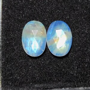 May include: Two oval-shaped, faceted moonstone gemstones. The stones have a translucent, milky blue color with iridescent flashes of yellow and white. The gemstones are displayed on a dark, textured surface.