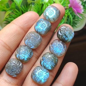 May include: A selection of carved labradorite gemstones. Each square-shaped stone features a floral design with blue and grey hues. The gemstones are held in a hand, with a blurred green and pink background.