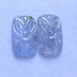 May include: Two pale purple, rectangular gemstones with carved floral designs. Each stone features a central flower with surrounding leaf patterns. The gemstones are set against a white background, showcasing their intricate details.