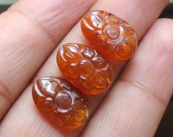 May include: Three oval-shaped, amber-colored gemstones with carved floral designs. The gemstones are held in a hand, showcasing their polished surfaces and intricate details. The carvings feature a central circular element surrounded by petal-like shapes.