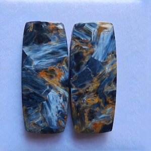 May include: Two rectangular Pietersite gemstones with a polished finish. The stones display a swirling pattern of deep blue, light blue, and golden-orange hues. The gemstones are set against a light blue background.