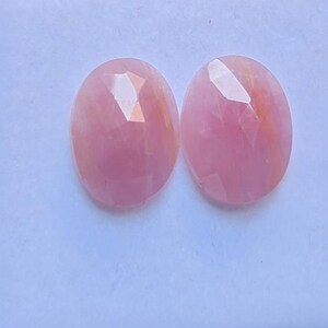 Peut inclure: Deux pierres précieuses roses de forme ovale et à facettes avec une surface lisse et polie. Chaque pierre présente un aspect translucide avec de subtiles variations d'intensité de couleur, incluant des teintes rose clair et des traînées orange.
