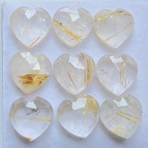 May include: Nine heart-shaped rutilated quartz gemstones arranged in a three-by-three grid. Each clear, faceted stone features unique golden needle-like inclusions embedded within the crystalline structure, creating a distinct pattern in every piece.