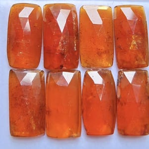 May include: Eight rectangular, faceted orange gemstones arranged in two rows of four. Each stone features a polished surface with geometric cuts that reflect light, displaying varying shades of translucent amber and deep orange hues throughout.