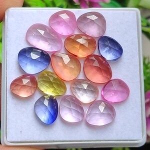 Puede incluir: Colección de gemas facetadas en varios colores, incluyendo rosa, naranja, amarillo y azul, dispuestas en una bandeja blanca. Las gemas tienen una superficie pulida y reflectante, y son de diferentes formas y tamaños.