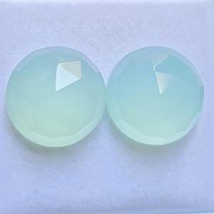 Peut inclure: Deux pierres précieuses rondes et facettées d'une couleur vert menthe pâle et translucide. Chaque pierre présente une surface lisse et polie avec des facettes géométriques sur les bords qui captent la lumière, créant un reflet subtil.