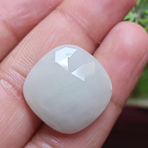 Puede incluir: Una piedra preciosa blanca translúcida de talla cojín con una superficie lisa y pulida y una sección superior facetada. La piedra tiene una forma cuadrada redondeada con bordes suaves, mostrando un aspecto lechoso y semiopaco.