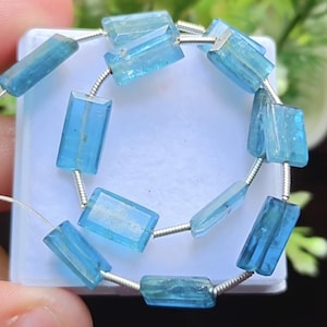 May include: A strand of rectangular, light blue gemstones strung on a silver wire. The gemstones are translucent and have a polished appearance. The beads are displayed against a light blue background.