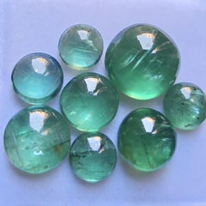 May include: A collection of eight round, smooth, translucent green gemstones of varying sizes. Each stone features a polished, domed cabochon cut with visible internal inclusions and light reflections on the curved surfaces, displaying shades of mint and emerald green.
