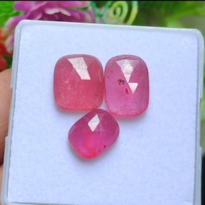 Op de afbeelding: Drie gefacetteerde, kussenvormige edelstenen in verschillende roze tinten worden gepresenteerd op een wit oppervlak. De edelstenen hebben een gepolijste uitstraling en reflecteren licht, waardoor hun interne structuur wordt getoond. De opstelling suggereert een verzameling edelstenen.