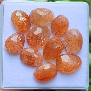 May include: A collection of ten faceted sunstone gemstones. The stones are oval-shaped with a warm orange hue and sparkling inclusions. They are displayed in a white tray, showcasing their polished surfaces and natural beauty. Ideal for jewelry making.