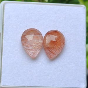 May include: Two teardrop-shaped gemstones with a faceted cut. The gems have a warm, reddish-brown color with internal inclusions. They are displayed in a white, square container, suggesting they are for jewelry making or collecting.