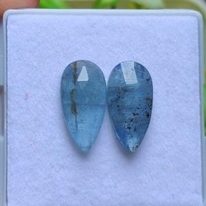 Pode incluir: Duas pedras preciosas de água-marinha facetadas em forma de pêra. As pedras preciosas são de cor azul clara com inclusões internas. São exibidas em uma superfície branca dentro de um recipiente transparente.