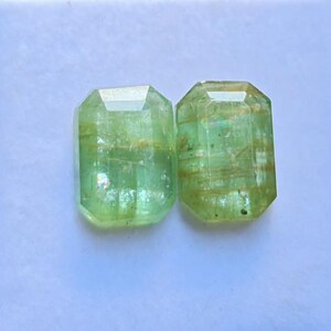 May include: Two light green, rectangular-cut gemstones with multiple facets. The gems have a transparent appearance with internal inclusions and a polished surface. The stones are displayed on a white background.
